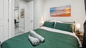 A hotel room with a bed, green bedding, a painting of a beach, and a nightstand with a lamp. - 2BR Basement Suite + Sofa Bed | Near Airport (Calgary)