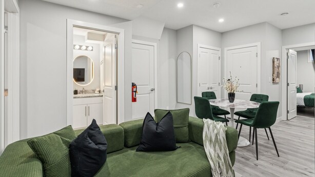 A modern living room with a green sofa, a dining table with chairs, and a white door with lights. - 2BR Basement Suite + Sofa Bed | Near Airport (Calgary)