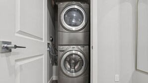 A compact laundry room with two stacked front-loading washing machines and a door with a handle. - 2BR Basement Suite + Sofa Bed | Near Airport (Calgary)