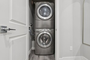 A compact laundry room with two stacked front-loading washing machines and a door with a handle. - 2BR Basement Suite + Sofa Bed | Near Airport (Calgary)