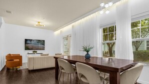 A dining area with a wooden table, four chairs, a sofa, and a television on the wall. - Thematic 4 bdr House Near Disney - 11-105lv (Kissimmee)