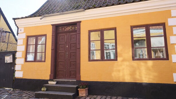 A yellow building with a brown door and windows, a tiled roof, and a cobblestone ground.