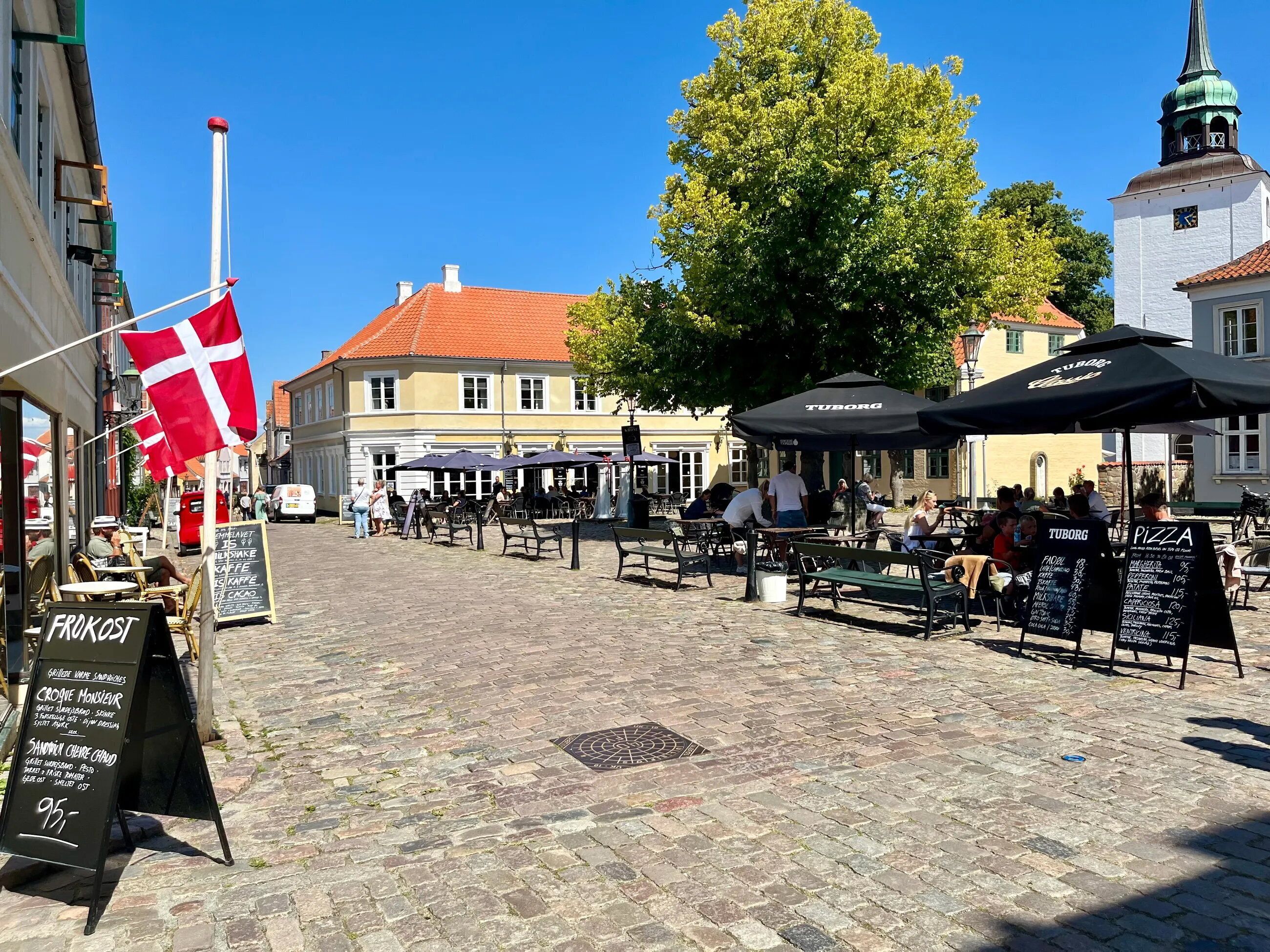 Une rue pavée avec des tables et des chaises installées à l’extérieur, un drapeau et un bâtiment surmonté d’une tour horloge.