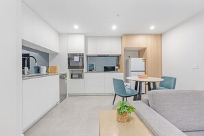 A modern kitchen with white cabinets, a microwave, and a sink. There is a dining area with a table and chairs, and a grey sofa.