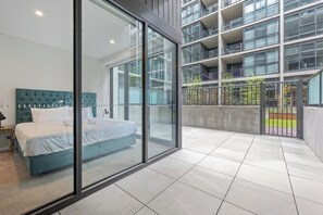 A modern hotel room with a large bed, a glass wall, and a view of a multi-story building.