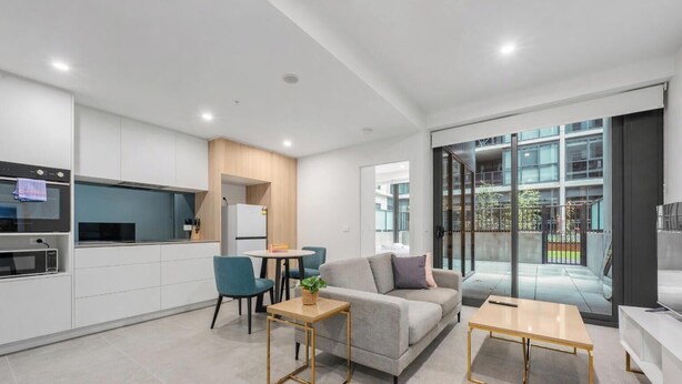 Un appartement moderne doté d’une kitchenette, d’un canapé gris, d’une petite table à manger et d’une grande porte-fenêtre coulissante donnant sur un espace extérieur.