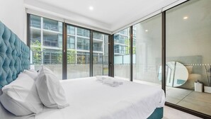 Une chambre moderne avec un grand lit, une vue sur un immeuble par la fenêtre et une porte-fenêtre coulissante donnant sur un balcon.