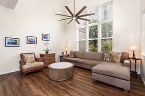 Un salon comprenant un canapé d’angle, une table basse ronde, un fauteuil en osier, un ventilateur de plafond et trois tableaux encadrés accrochés au mur.