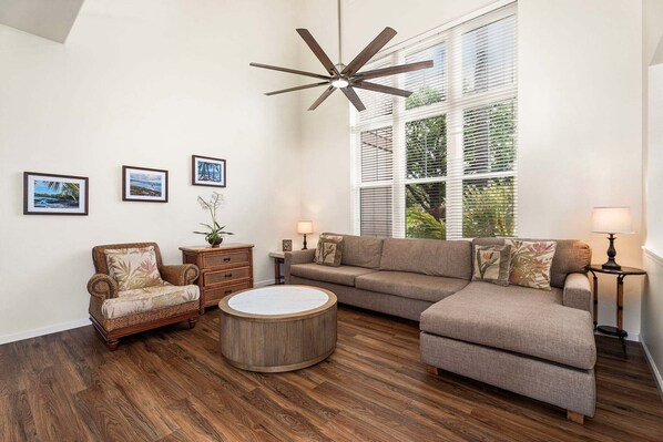 Ein Wohnzimmer mit einem Ecksofa, einem runden Couchtisch, einem Korbstuhl, einem Deckenventilator und drei gerahmten Bildern an der Wand.