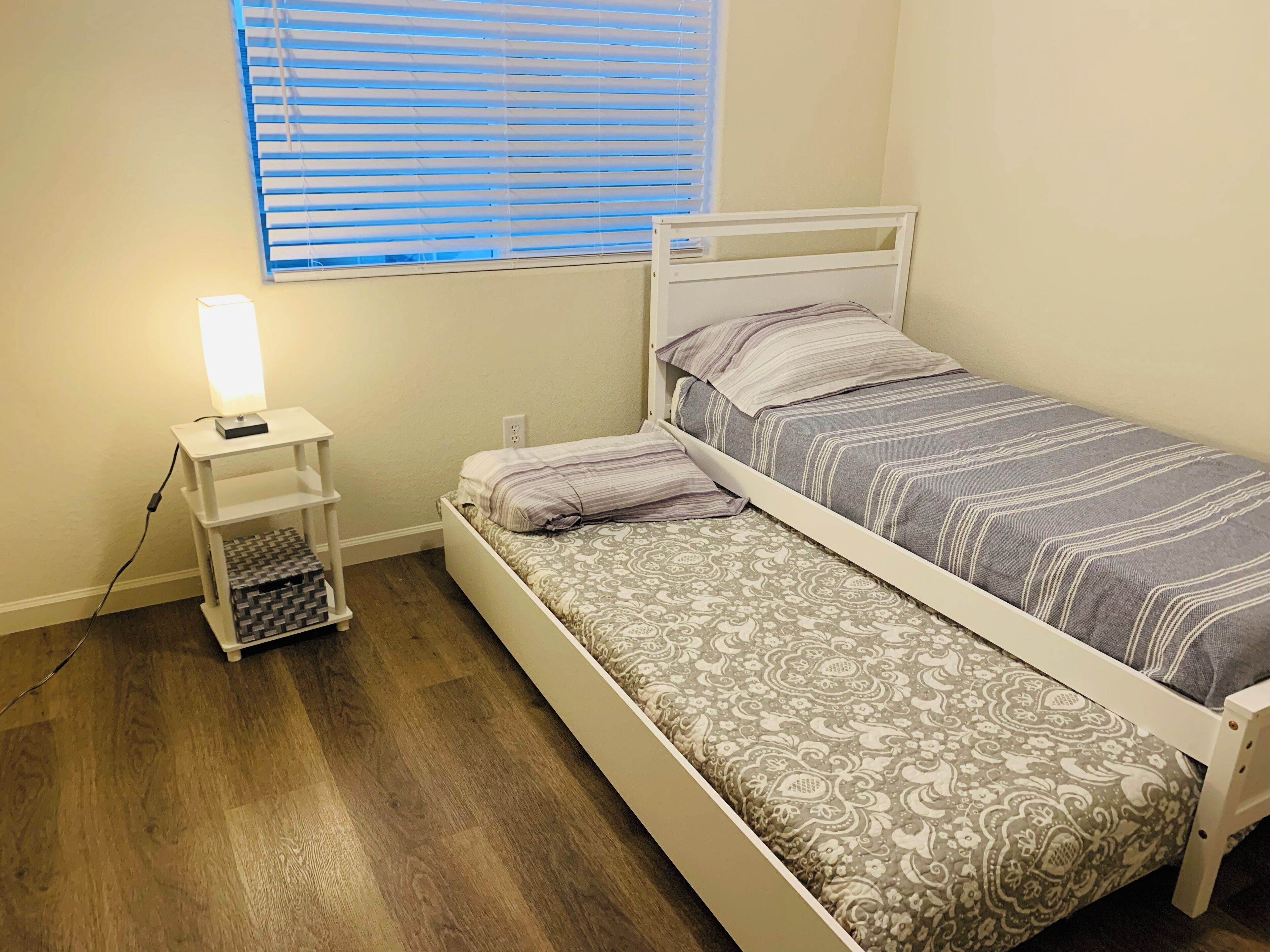 A small, neatly arranged bedroom with a single bed, a bedside table with a lamp, and a window with blinds.