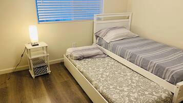 A small, neatly arranged bedroom with a single bed, a bedside table with a lamp, and a window with blinds.