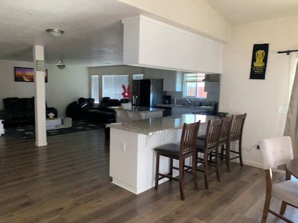 Una cocina moderna con una isla central y taburetes, un comedor con mesa y sillas, y una sala de estar con sofá y una mesa de centro.