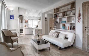 Un salon avec un canapé blanc, une table basse et un fauteuil. Au fond, on aperçoit une cuisine avec des meubles blancs et une fen être.