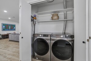 A laundry room with two stainless steel front-loading washers and dryers, a white shelf with a woven basket, and a painting on the wall. - Fabulous 5 Bedroom Elegance Near LSU -Single Level home with Mother-in-Law suite (Baton Rouge)