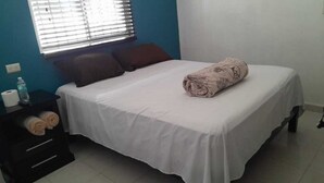 Un lit bien fait, recouvert d’une couverture blanche, avec une serviette enroulée posée sur le dessus. Il y a une fenêtre avec des stores, une table de chevet sur laquelle se trouvent une bouteille d’eau et une boîte de mouchoirs, ainsi qu’une commode de couleur sombre dotée de deux tiroirs.