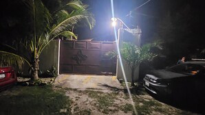 Une entrée fermée par un portail, avec un chemin en béton menant à un bâtiment, le tout entouré de palmiers et avec une voiture rouge garée sur le côté gauche.