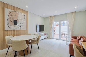 A modern living room with a round dining table, beige chairs, a flat-screen TV, a white cabinet, and a large abstract painting.
