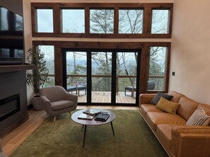 A spacious living room with a large window, a fireplace, a sofa, and a coffee table. - Modern home with Mountain Views, Hot Tub, EV outlet, Family and Dog friendly, (Mineral Bluff)