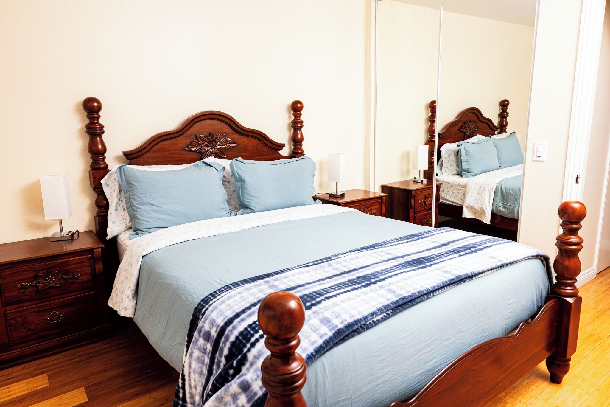 Een keurig opgemaakt bed met een houten hoofdbord, een nachtkastje met een lamp en een spiegel aan de muur.