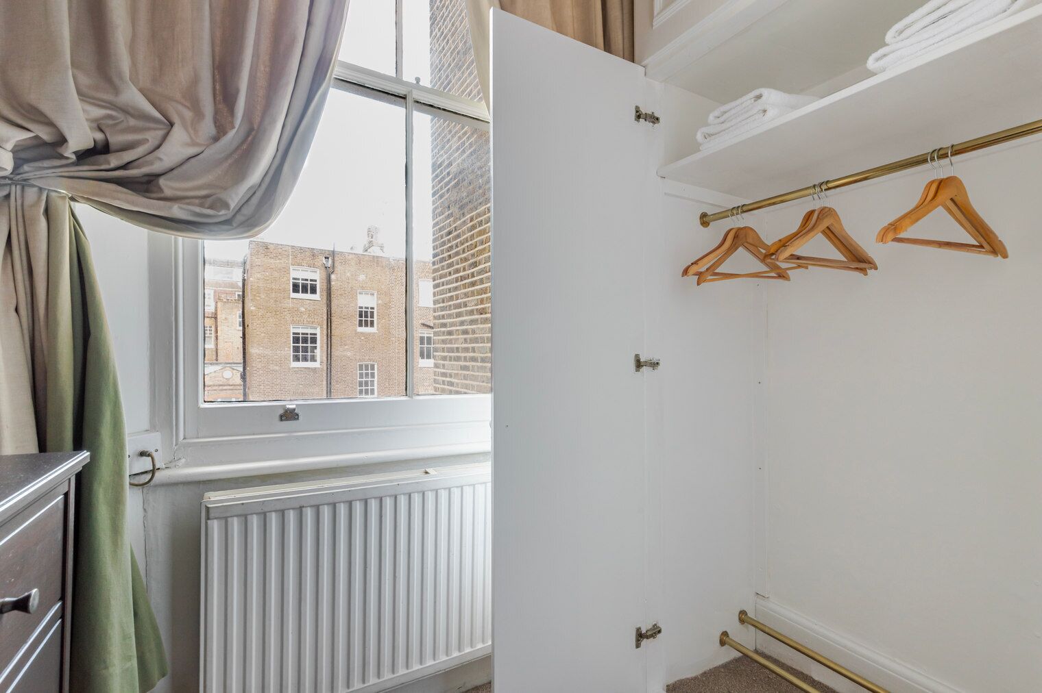 Une pièce avec un radiateur, une fenêtre avec des rideaux, un porte-manteau en bois et une vue sur un bâtiment en briques à l’extérieur.