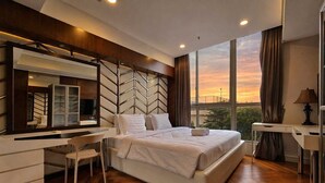 A modern bedroom with a large bed, a desk, a chair, a mirror, and a view of the sunset through large windows. - Summit By SanLiving - 2BR (Jakarta)