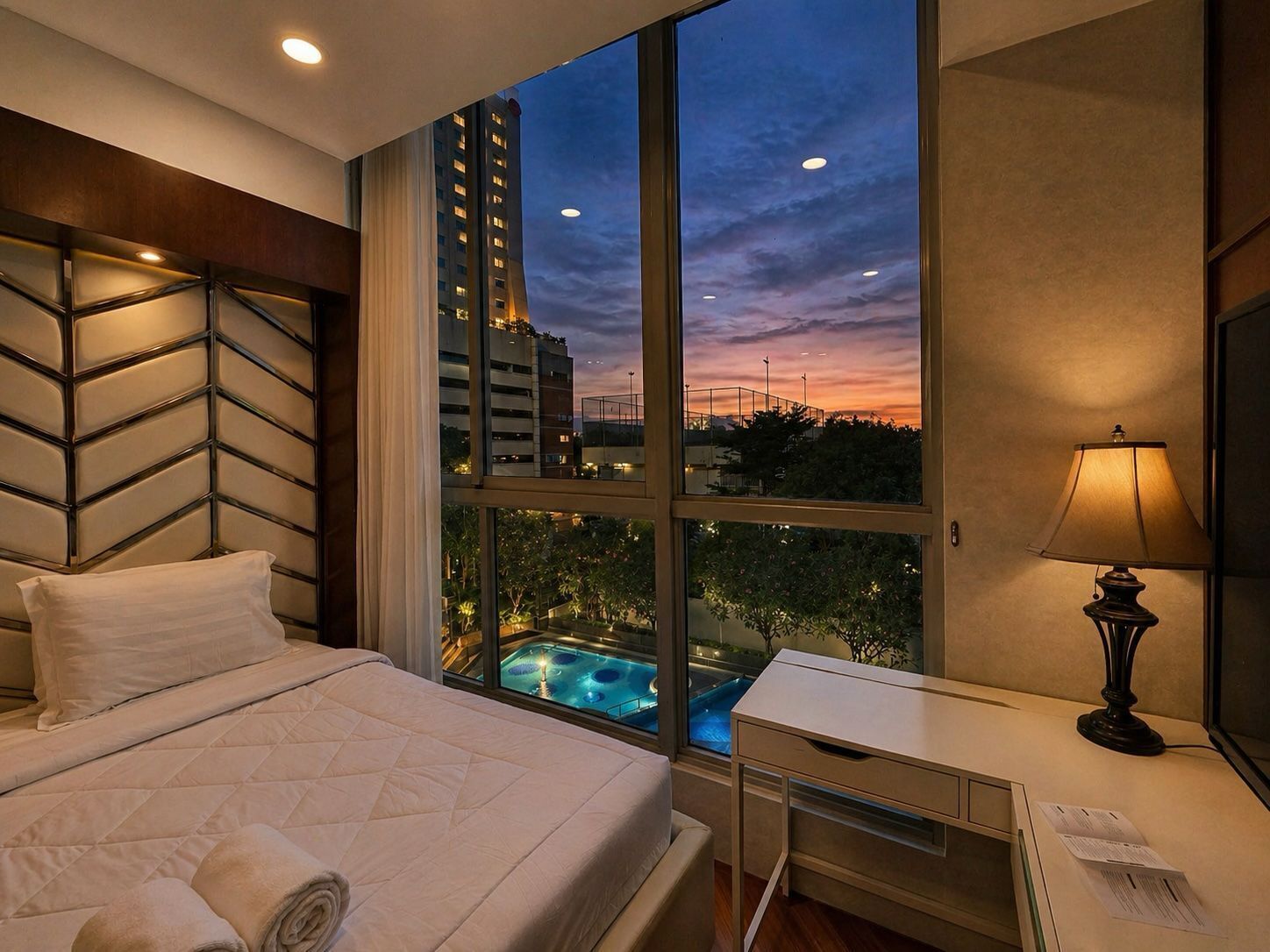 A hotel room with a bed, a desk, a lamp, and a large window with a view of a pool and buildings.
