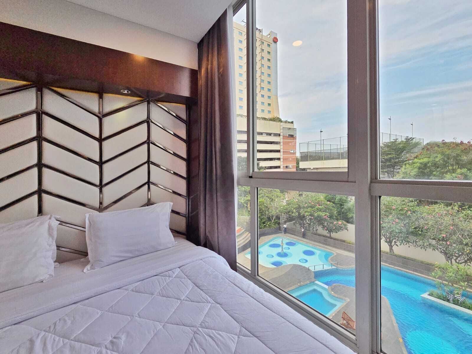 Hotelkamer met een groot raam, een bed met wit beddengoed, een hoofdeinde met een zigzaagpatroon en uitzicht op een zwembad en gebouwen.