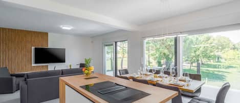 A modern kitchen with a central island, dining area with chairs, a TV mounted on the wall, and large windows offering a view of the outdoors.
