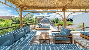 Un espacio exterior techado con techo de bambú, asientos acolchados y vistas a la vegetación circundante.