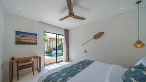 Un dormitorio con cama, escritorio, ventilador de techo, ventana grande con vista a una piscina y una hoja decorativa en la pared.