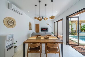A modern dining area with a wooden table, two wooden chairs, a water dispenser, and a TV mounted on the wall. - Villa Dara (Canggu)