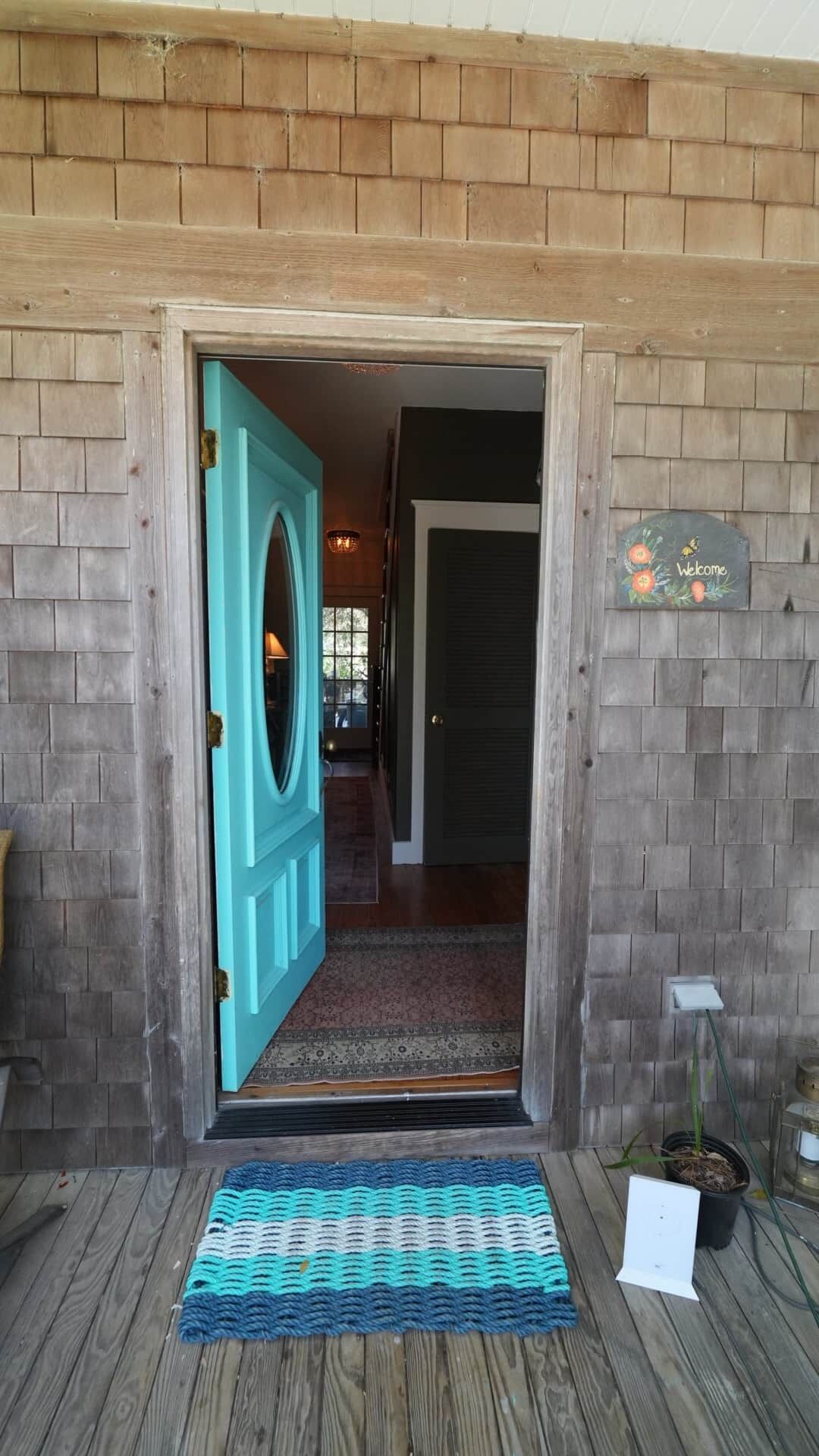 A wooden door with a teal frame leading to a hallway with a patterned rug and a welcome sign.