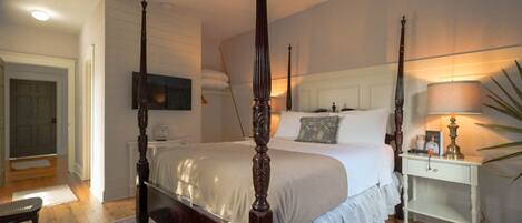 A four-poster bed with a white bedspread, a bedside table with a lamp, and a large plant on the right.