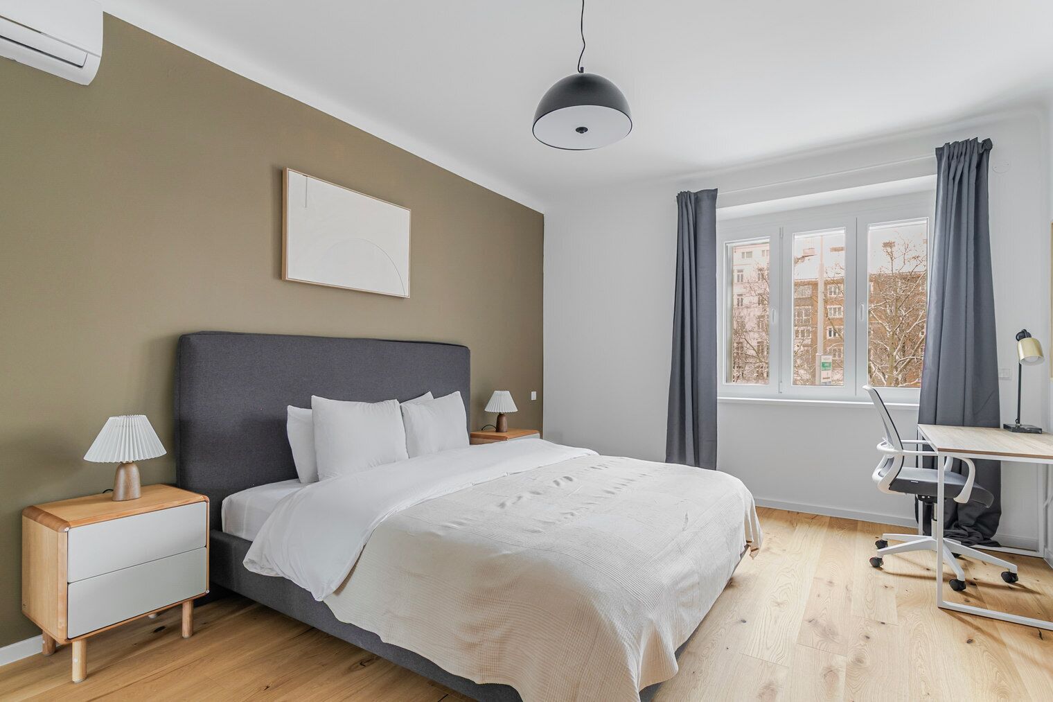A modern hotel room with a large bed, bedside tables with lamps, a desk with a chair, and a window with curtains.