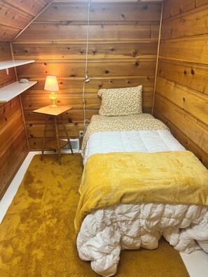Una habitación pequeña y acogedora con una cama, una mesita de noche con lámpara y una pared de madera.