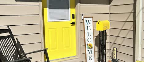 A house with a yellow door, a welcome sign, a mailbox, and a bench outside.