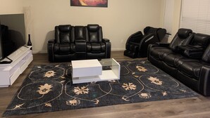 Ein Wohnzimmer mit einem schwarzen Ledersofa, einem weißen Couchtisch, einem schwarzen Massagesessel, einem Massagetisch, einem Flachbildfernseher und einem Gemälde, das einen Sonnenuntergang zeigt.