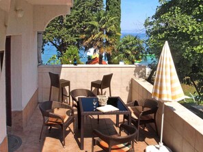 A terrace with wicker furniture, a table, and an umbrella, overlooking a pool and trees. - Holiday apartment Apartment Azzurra, first row to the sea (Njivice)