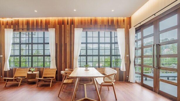 A spacious room with a wooden floor, a central table, and several wicker chairs. Large windows with curtains provide natural light and views of greenery outside. - Harley 1 BDR Condo Kata Hill, 3 Mins To kata Beach (Karon)