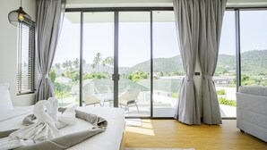 A hotel room with a bed, a sofa, and a view of palm trees and hills. - Camellia Pool Villa,Near Nai Harn And Rawai Beach (Tambon Rawai)