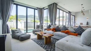 A spacious living room with a grey sofa set, a wooden coffee table, and a large window offering a view of the outdoors. - Camellia Pool Villa,Near Nai Harn And Rawai Beach (Tambon Rawai)