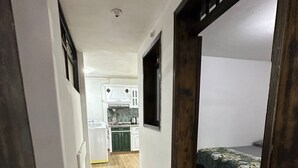 Un couloir étroit, doté d’un plancher en bois, mène à une chambre à coucher où se trouve un lit recouvert d’une couvre-lit à motifs verts. On aperçoit à l’arrière-plan un coin cuisine avec des armoires blanches et un fourneau.