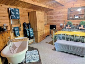 A wooden cabin interior with a bathtub, a bed with a patterned blanket, a wooden cabinet, and a mirror with a heart decoration.