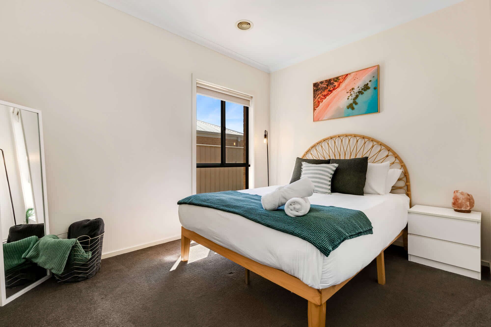 A neatly arranged bedroom with a bed, a bedside table, a wall art piece, and a view of the outside through a window.