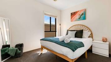 A neatly arranged bedroom with a bed, a bedside table, a wall art piece, and a view of the outside through a window.