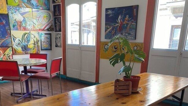 Un área de comedor con mesas y bancos de madera, una planta en maceta y coloridas obras de arte en las paredes.