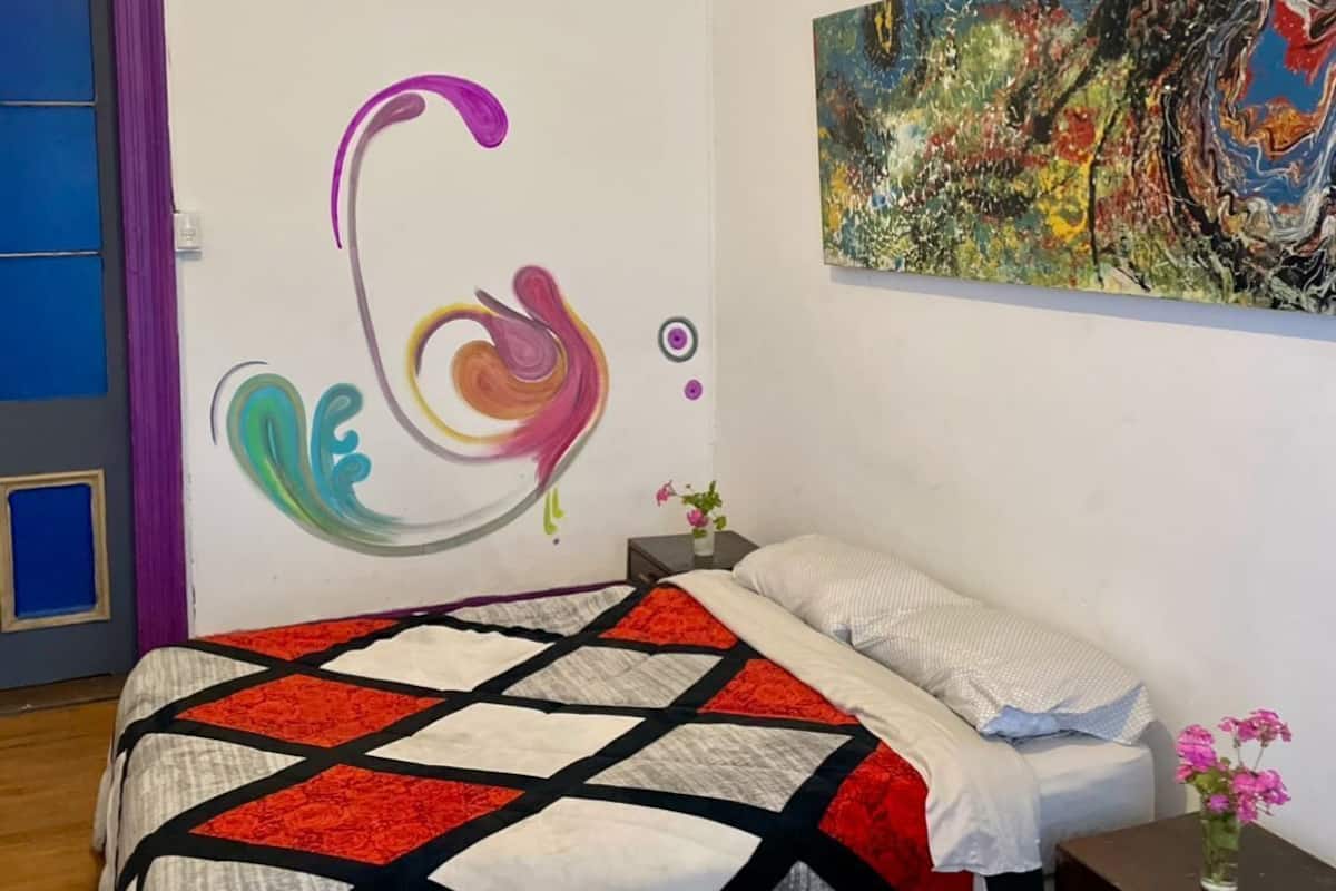 Une chambre à coucher avec un lit, un mur orné d’œuvres d’art abstraites, une porte et une table de chevet sur laquelle se trouve un vase.
