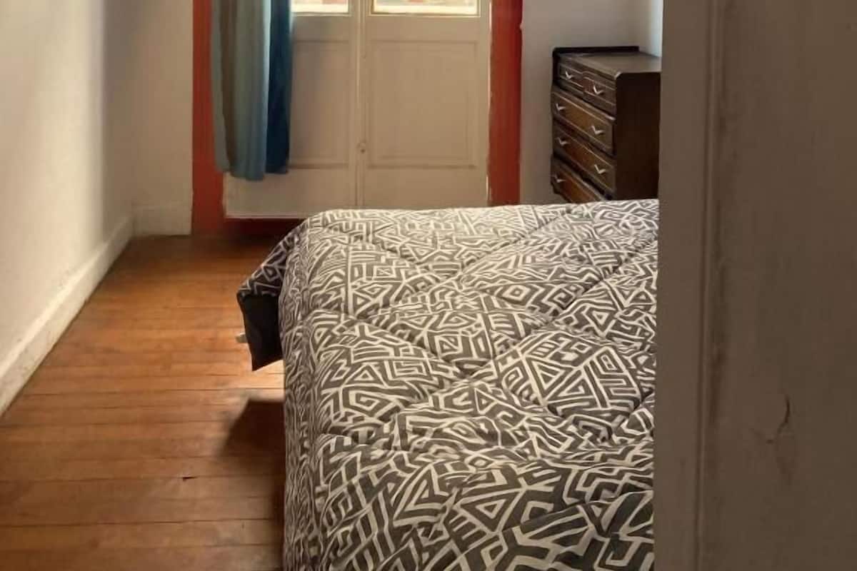Une chambre à coucher avec une couvre-lit à motifs, une commode en bois et une porte dotée d’un cadre rouge et de rideaux bleus.
