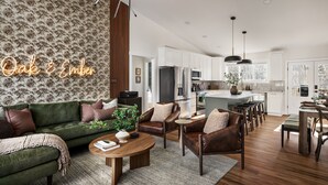 A modern living room with a green sofa, leather armchairs, and a wooden coffee table. A kitchen with white cabinets and a dining area with a wooden table and chairs is visible in the background.