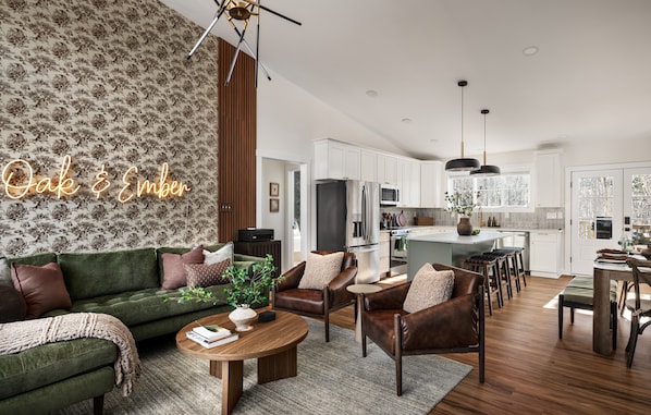A modern living room with a green sofa, leather armchairs, and a wooden coffee table. A kitchen with white cabinets and a dining area with a wooden table and chairs is visible in the background.